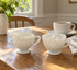 White milk glass bubble pattern creamer with cream and sugar bowl with sugar cubes on a wooden kitchen table beside a white coffee cup in morning light